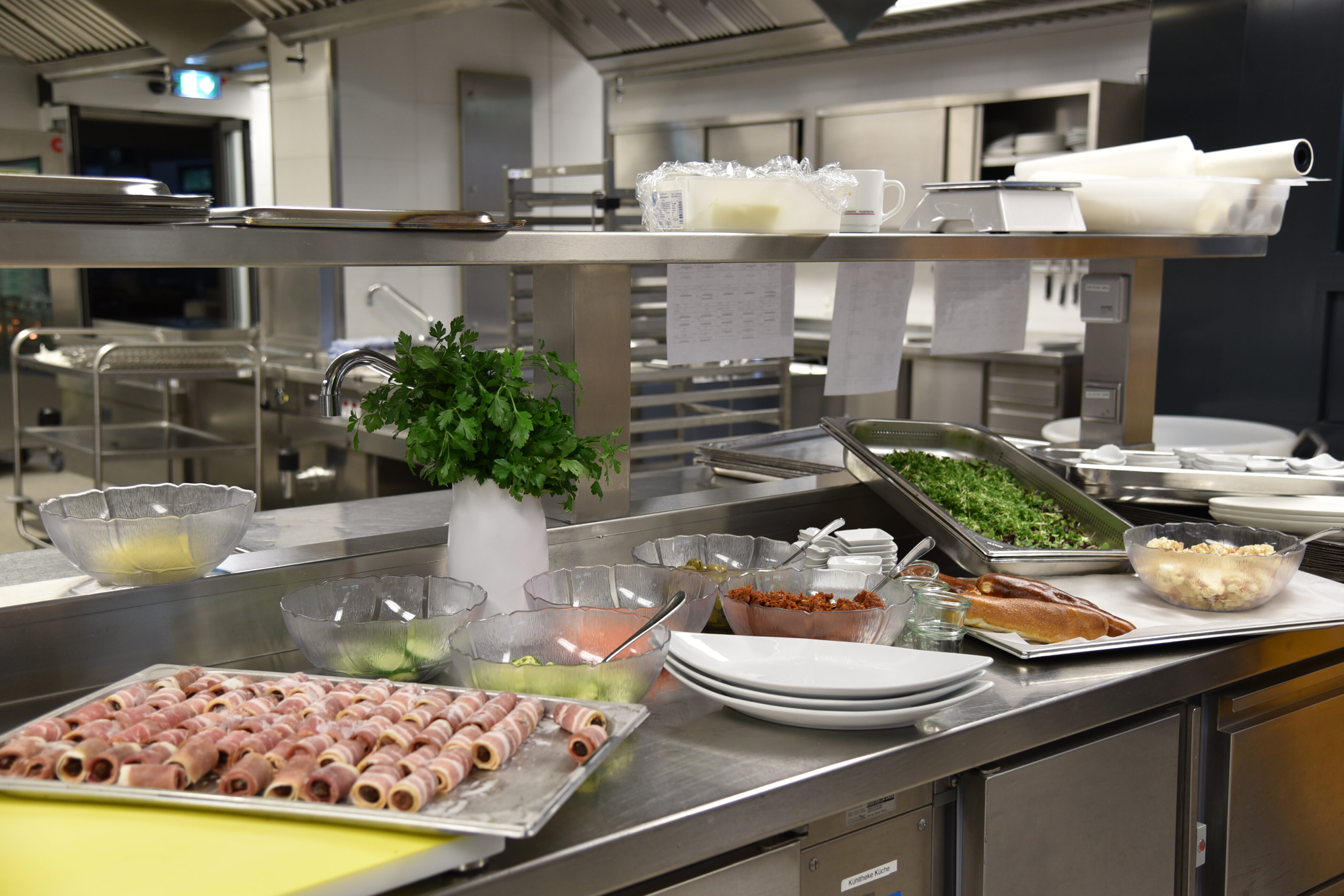 Ausschnitt einer Großküche mit MEtalleinrichtung, im Vordergrund einzelne Zutaten in Schüsseln, Tellern, Schinkenröllchen, Petersilie