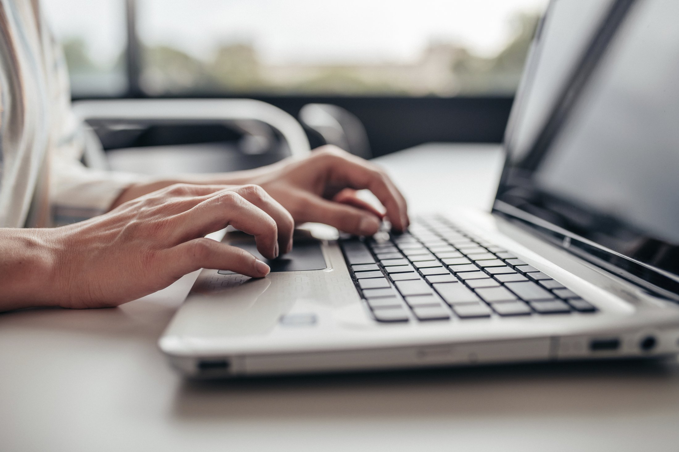 Eine Hand tippt auf der Tastatur eines Laptops. Im Hintergrund ist ein Fenster mit unscharfem Tageslicht zu sehen.
