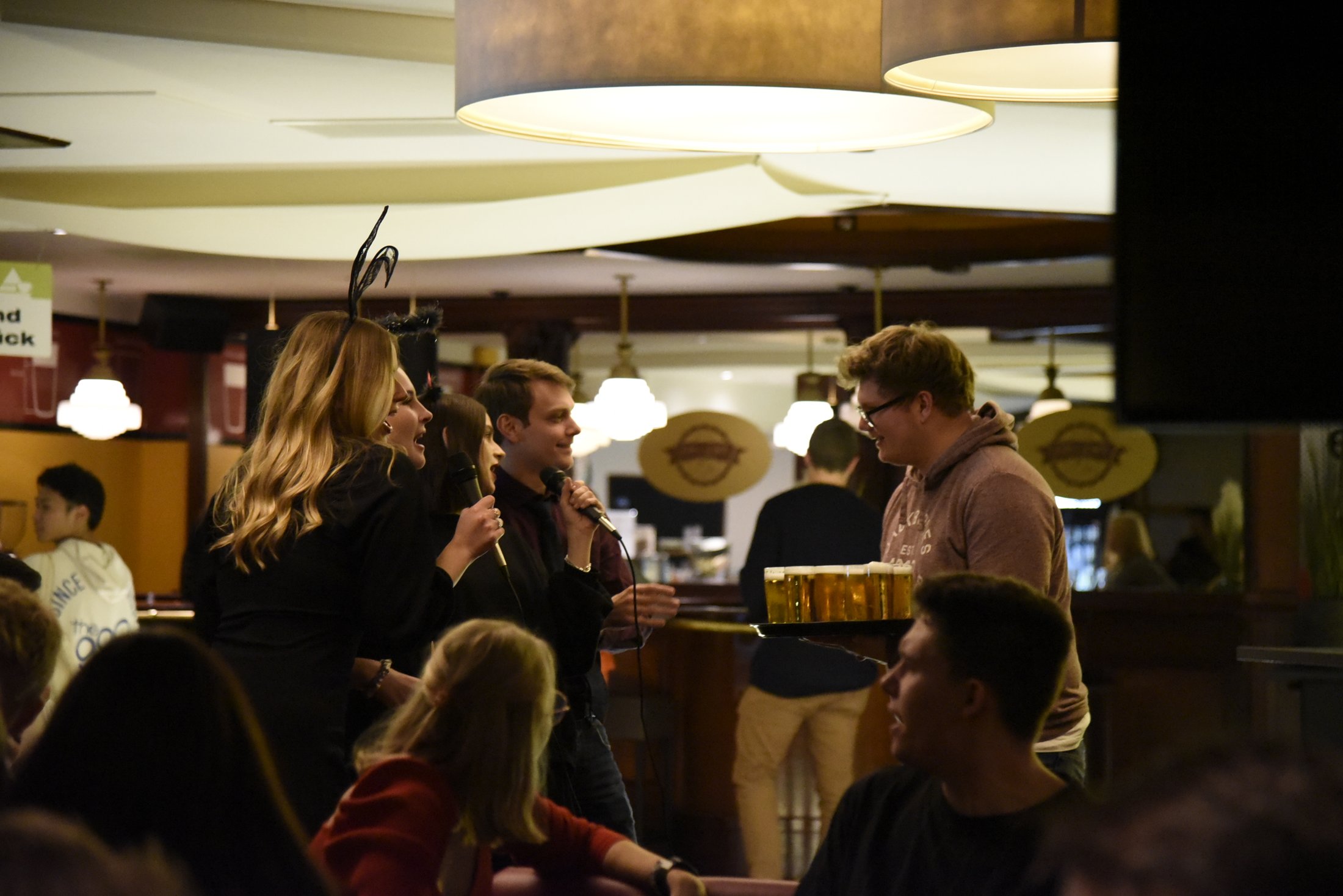 In einer Gastronomie-Szene unterhalten sich mehrere Personen, während ein Kellner mit einem Tablett voll Getränke serviert.