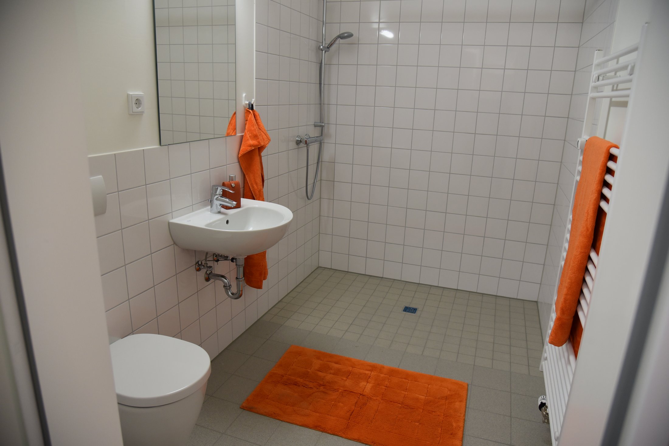 Barrierefreies Badezimmer im Studierendenwohnheim in Hamm, ausgestattet mit Dusche, Waschbecken und orangefarbenen Handtüchern.