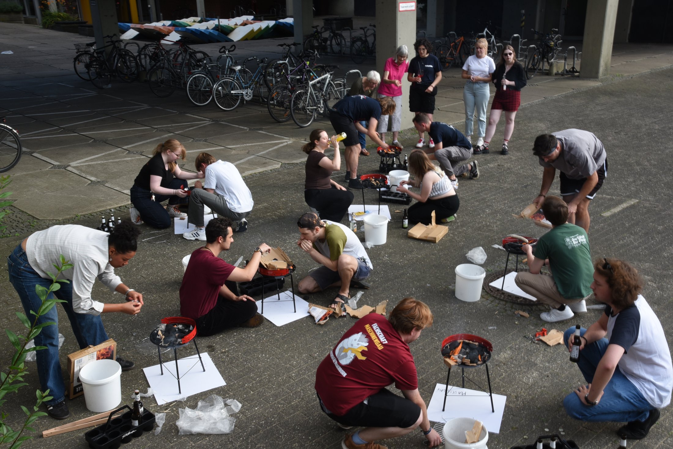 Gruppenevent im Freien mit mehreren Personen, die an kleinen Grillstationen arbeiten und zubereiten. Fahrräder im Hintergrund.