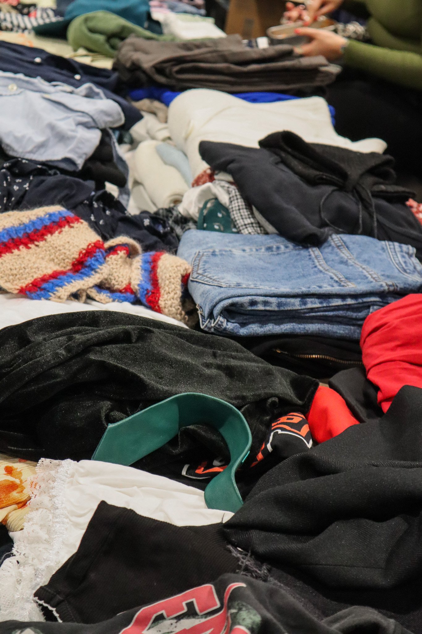 Vielfältige Kleidungstücke in verschiedenen Farben und Materialien liegen auf Tischen, einige sind gefaltet, andere übereinander.