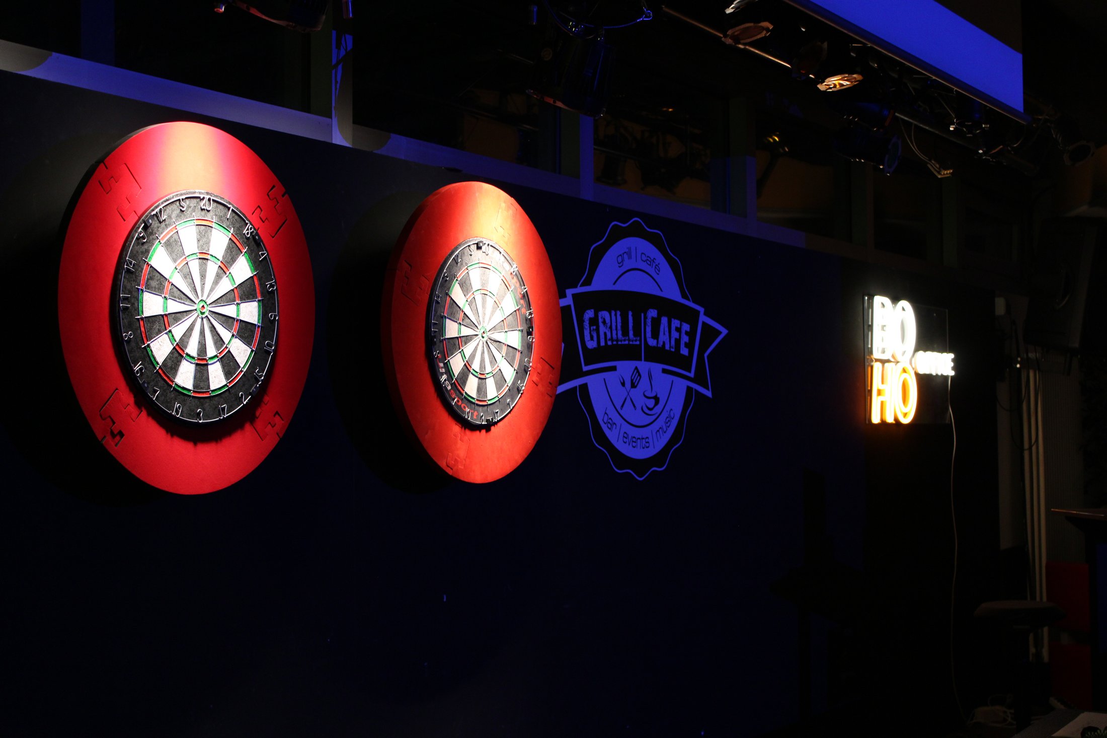 Zwei Dartscheiben an einer dunklen Wand, daneben ein leuchtendes Schild mit der Aufschrift „Grill Café“.