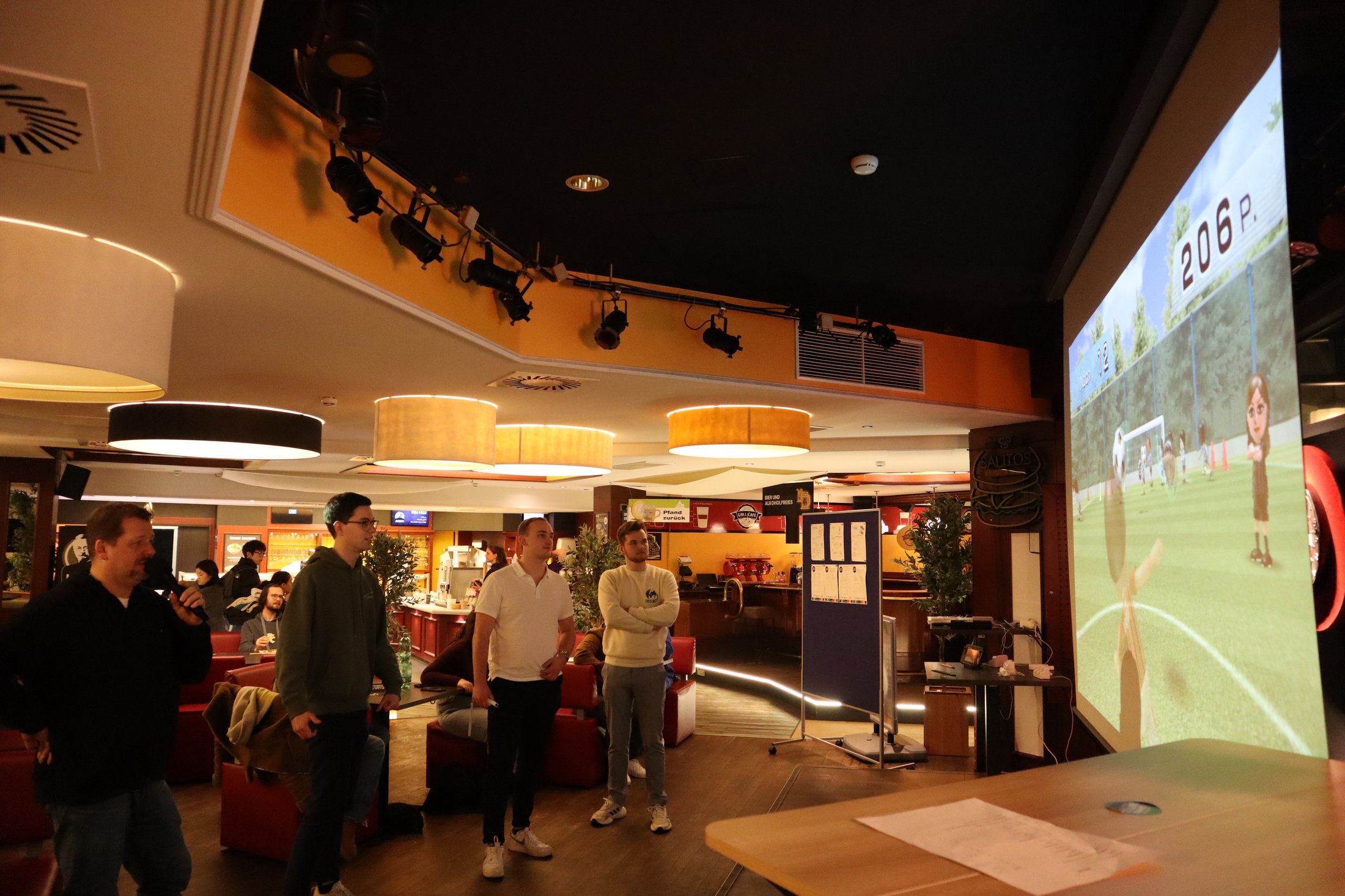 Gruppe von Personen im Interieur eines Lokals, die ein Spiel auf einer großen Leinwand verfolgen.