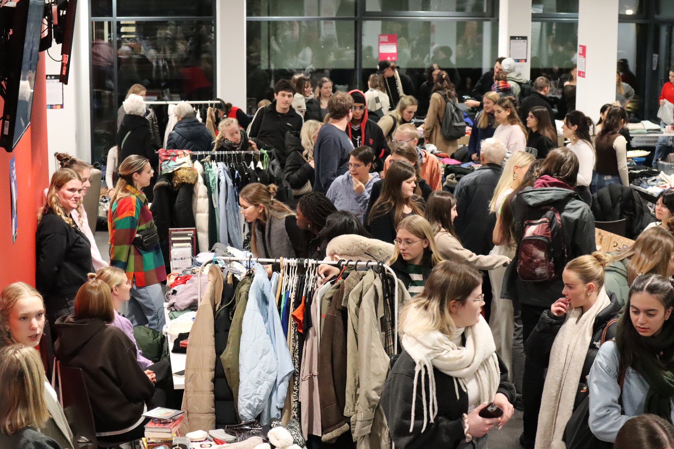 In einem Raum mit vielen Menschen, die an Ständen mit Kleidung stöbern. Verschiedene Textilien sind sichtbar.