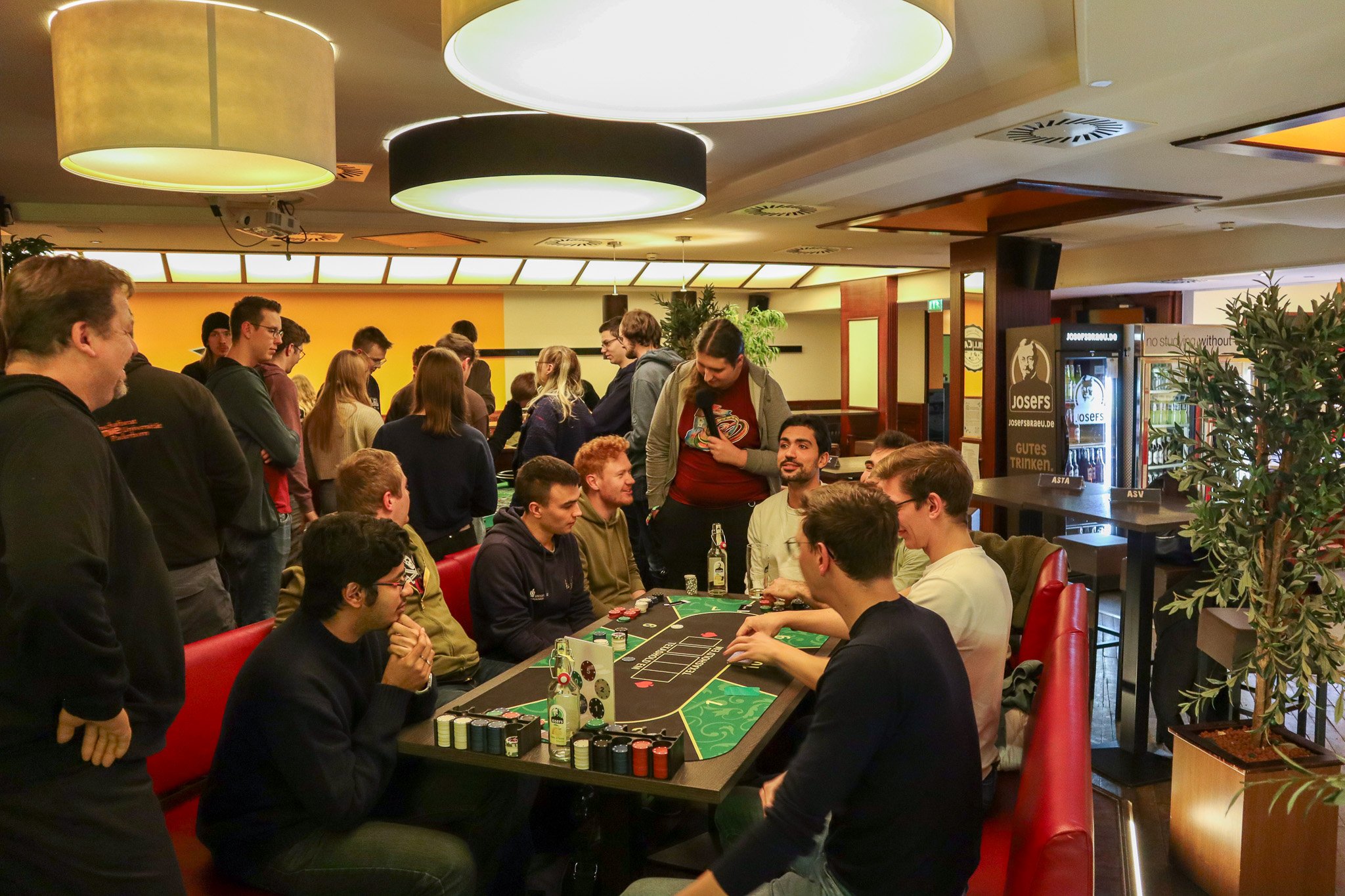 In einem Raum spielen Gruppen von Personen an Tischen Karten und unterhalten sich. Einige stehen, andere sitzen.