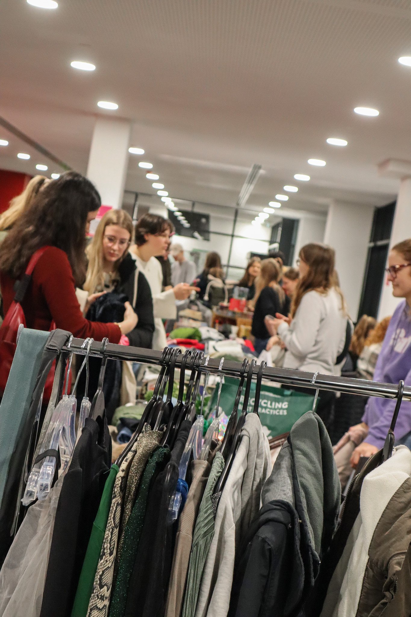 Menschen stehen in einem Raum und schauen sich Kleidung auf einem Verkaufsstand an. Im Hintergrund sind weitere Stände und Besucher.