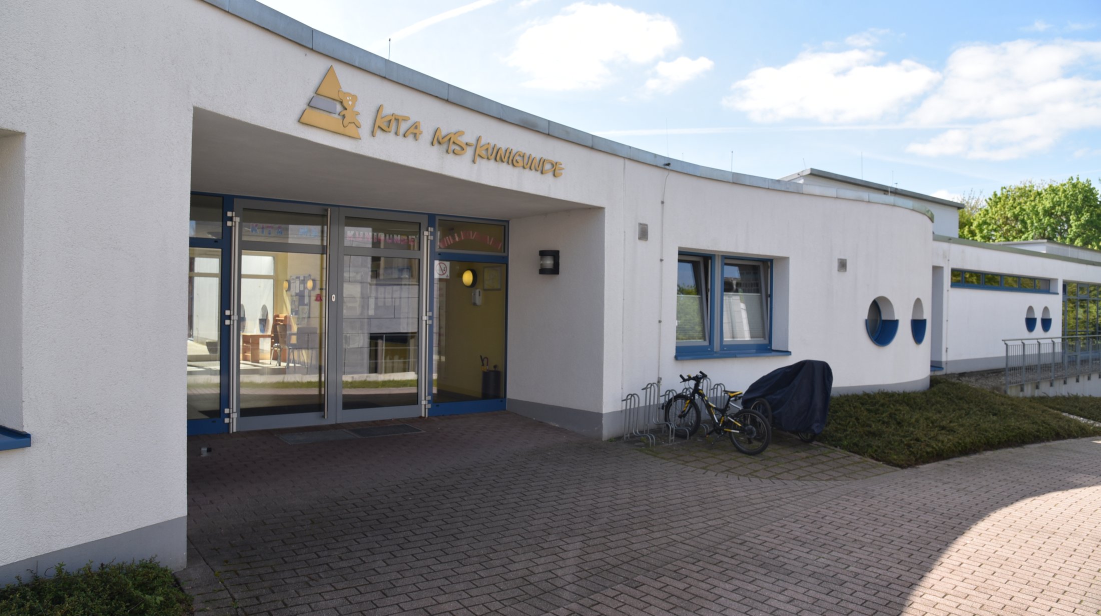 Eingang einer Kindertagesstätte mit großen Fenstern, bepflanztem Umfeld und Fahrradständer. Helle Fassadengestaltung.