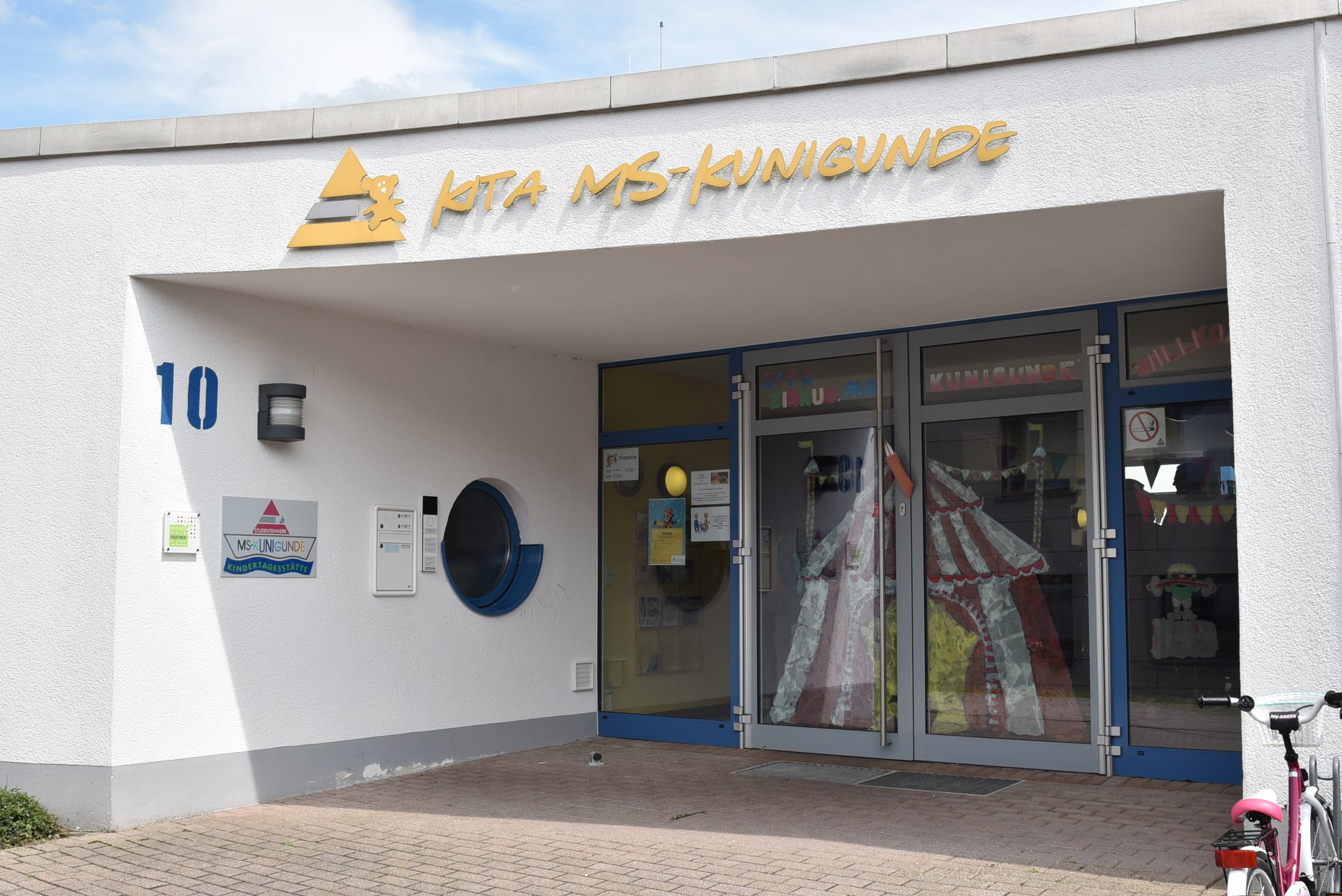 Eingangsbereich einer Kindertagesstätte mit bunten Fenstermalereien und einem Spielzeugfahrrad auf dem Weg.