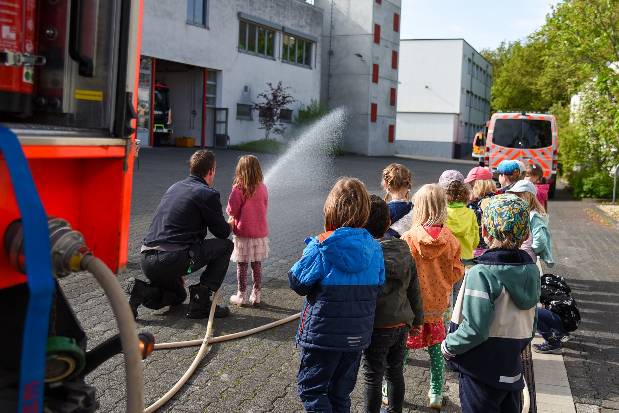 Kinder stehen in einer Reihe, während ein Feuerwehrmann einer kleinen Gruppe das Spritzen mit einem Feuerwehrschlauch zeigt.