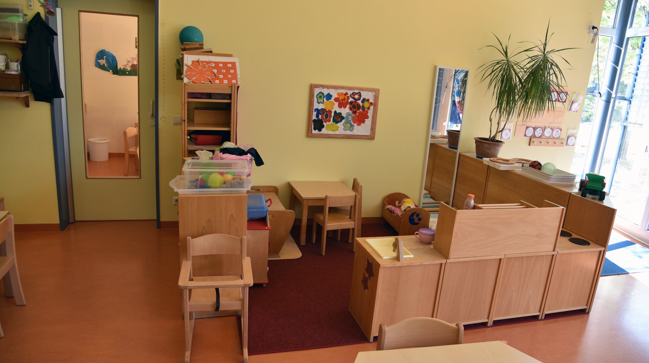 Heller Gruppenraum mit Tischen, Stühlen, Spielzeug und einer Pflanze. An der Wand hängt ein Kunstwerk.