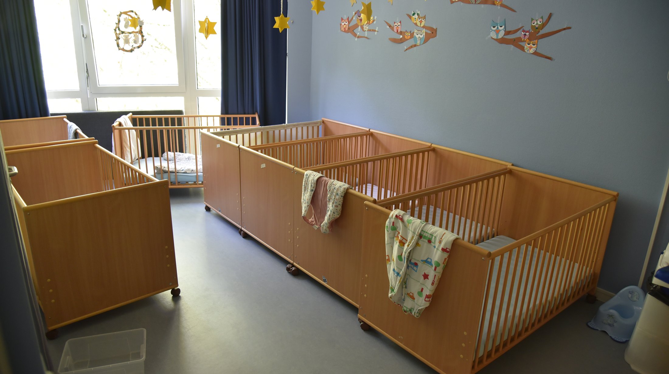 Raum mit mehreren Babybetten aus Holz, einer Fensterfront und einer Wanddekoration mit Eulen und Sternen.
