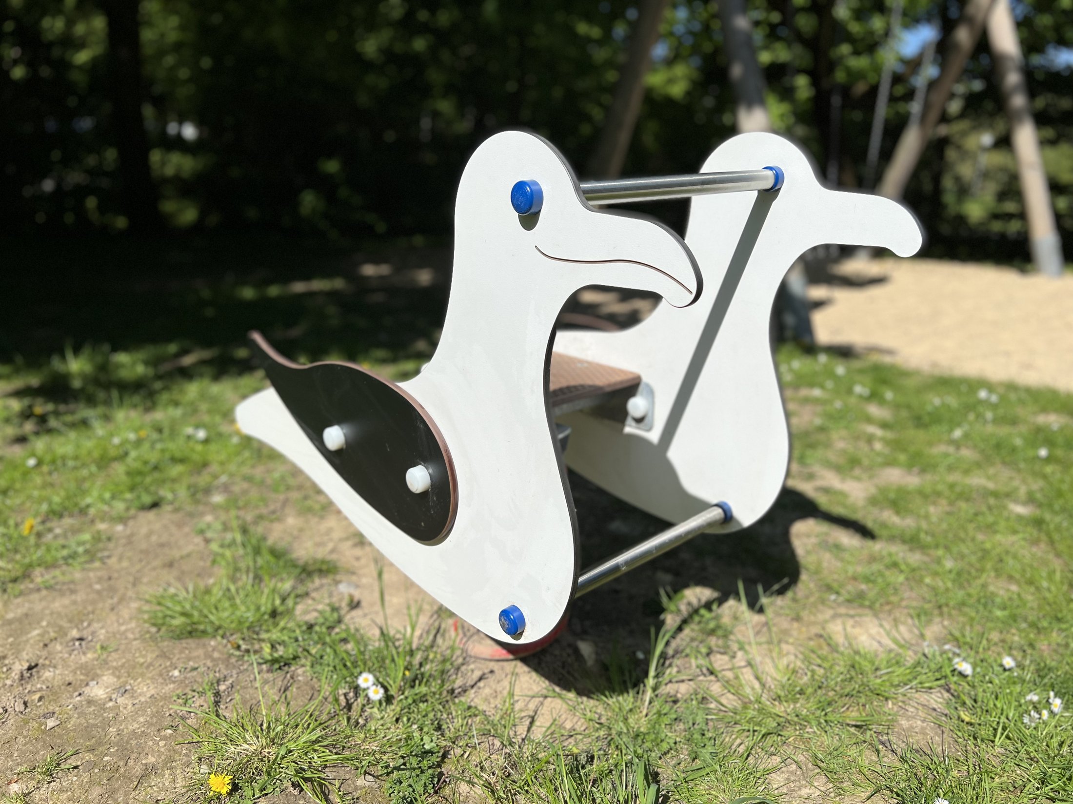 Ein hölzerner Wippelang mit Enten-Design steht auf einer grünen Wiese, umgeben von kleinen Blumen.