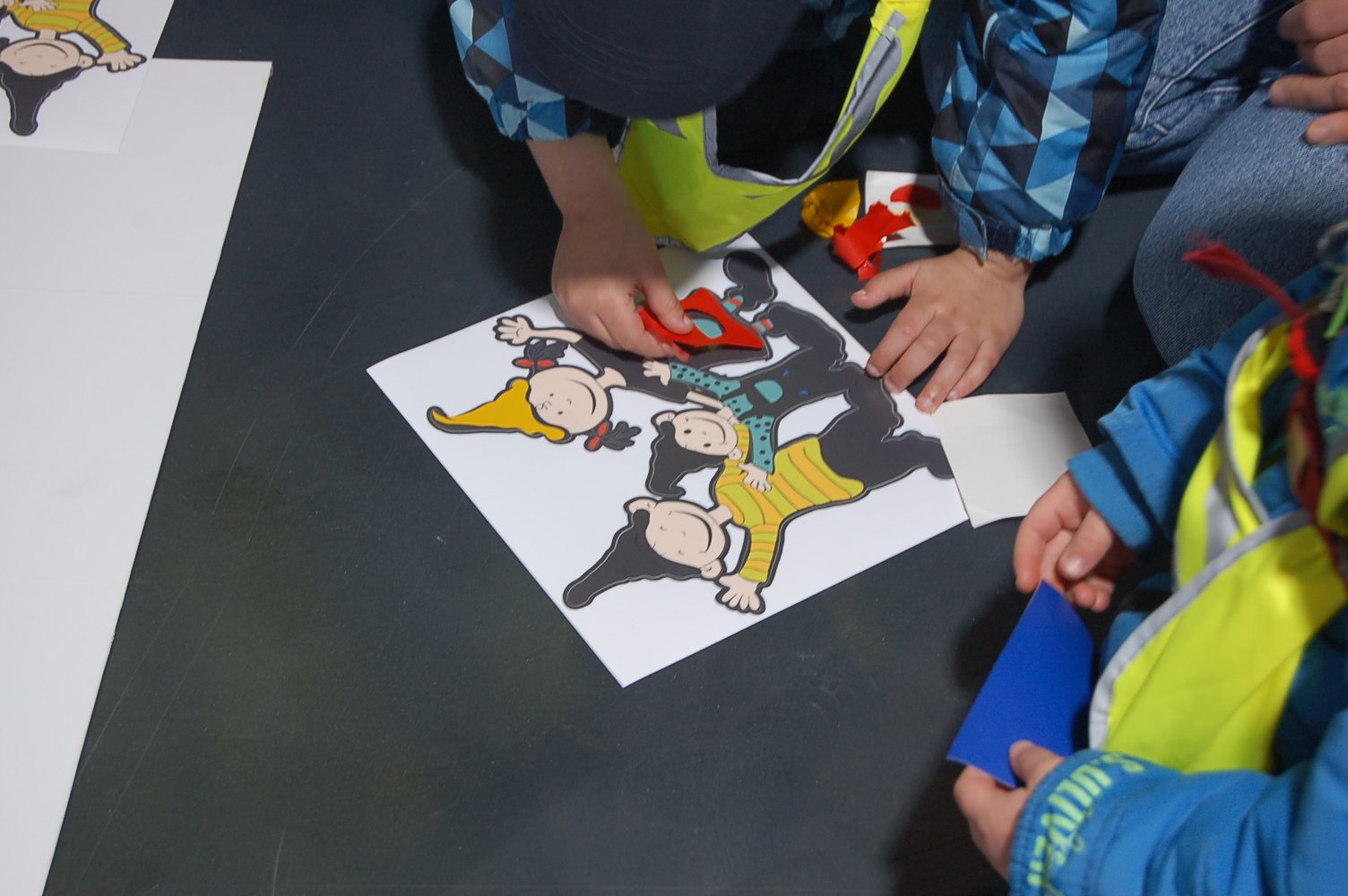 Kinder setzen farbige Formen auf ein Bild von drei fröhlichen Charakteren mit verschiedenen Hüten.