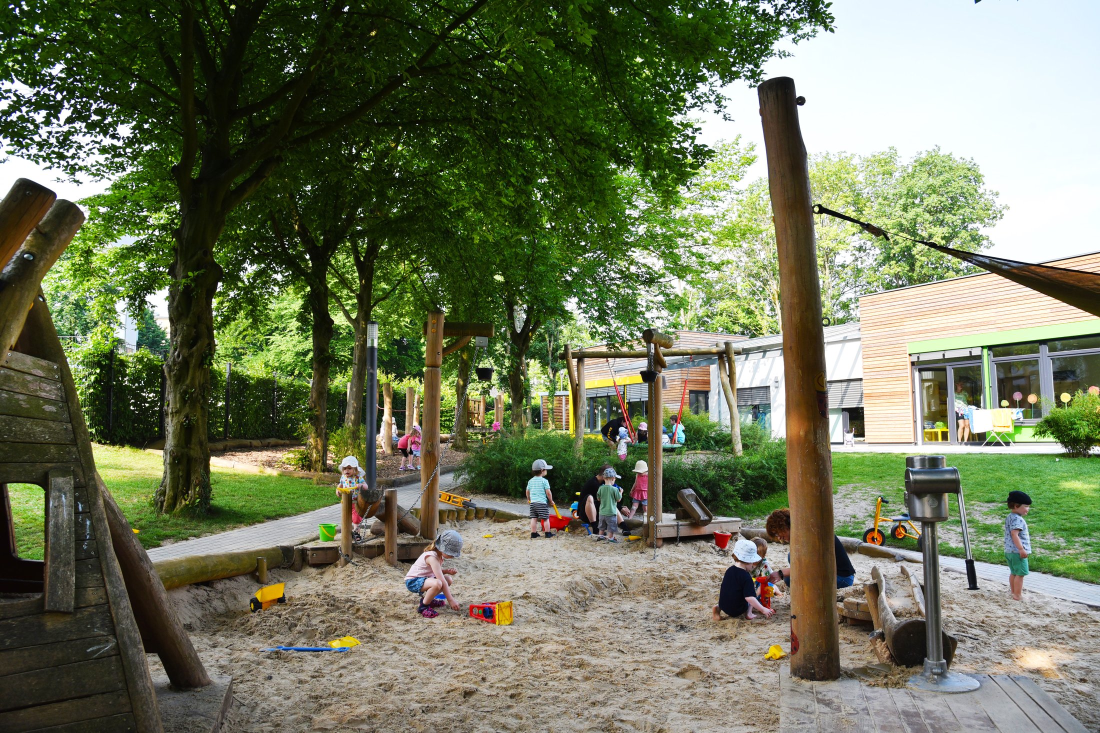 Spielplatz mit Sandkasten, Spielgeräten und Kindern, die im Freien spielen. Bäume und ein Gebäude im Hintergrund.