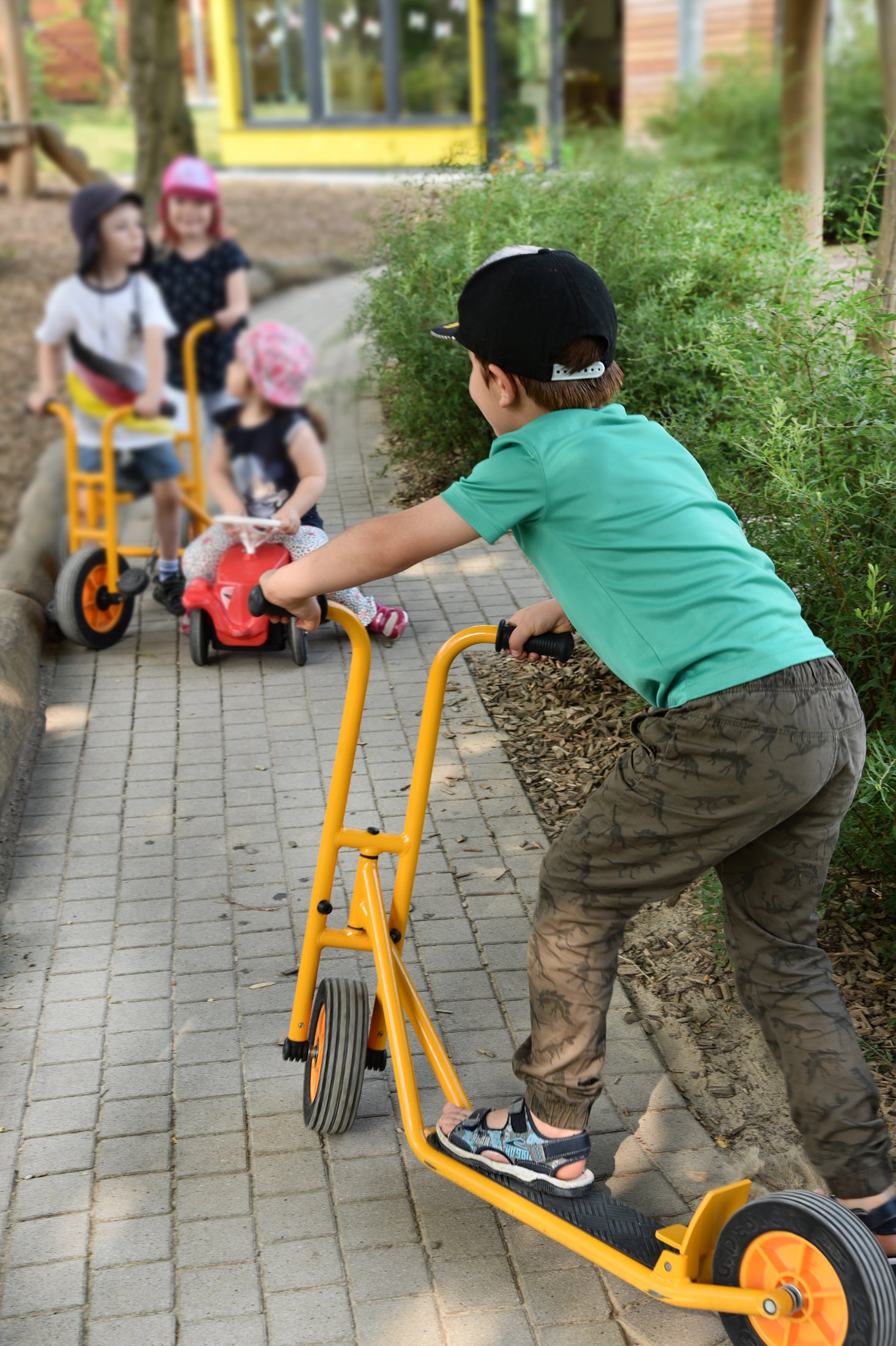 Kinder fahren auf einem gepflasterten Weg mit verschiedenen Fahrzeugen, umgeben von Grünflächen und einem Gebäude im Hintergrund.