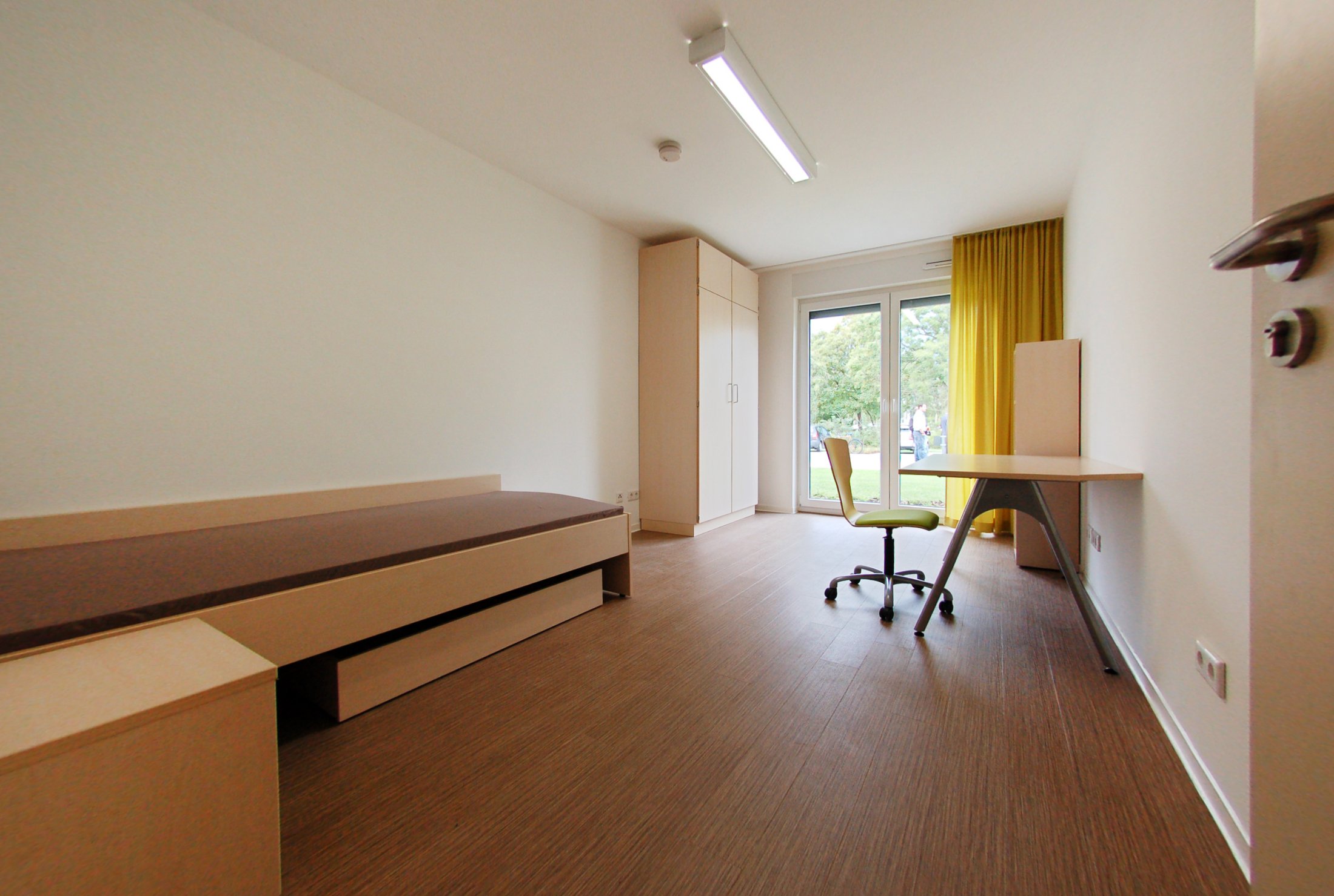 Studierendenwohnheim in Lippstadt: Helles Zimmer mit Bett, Schreibtisch, Stuhl und Fenster zur Gartenansicht.