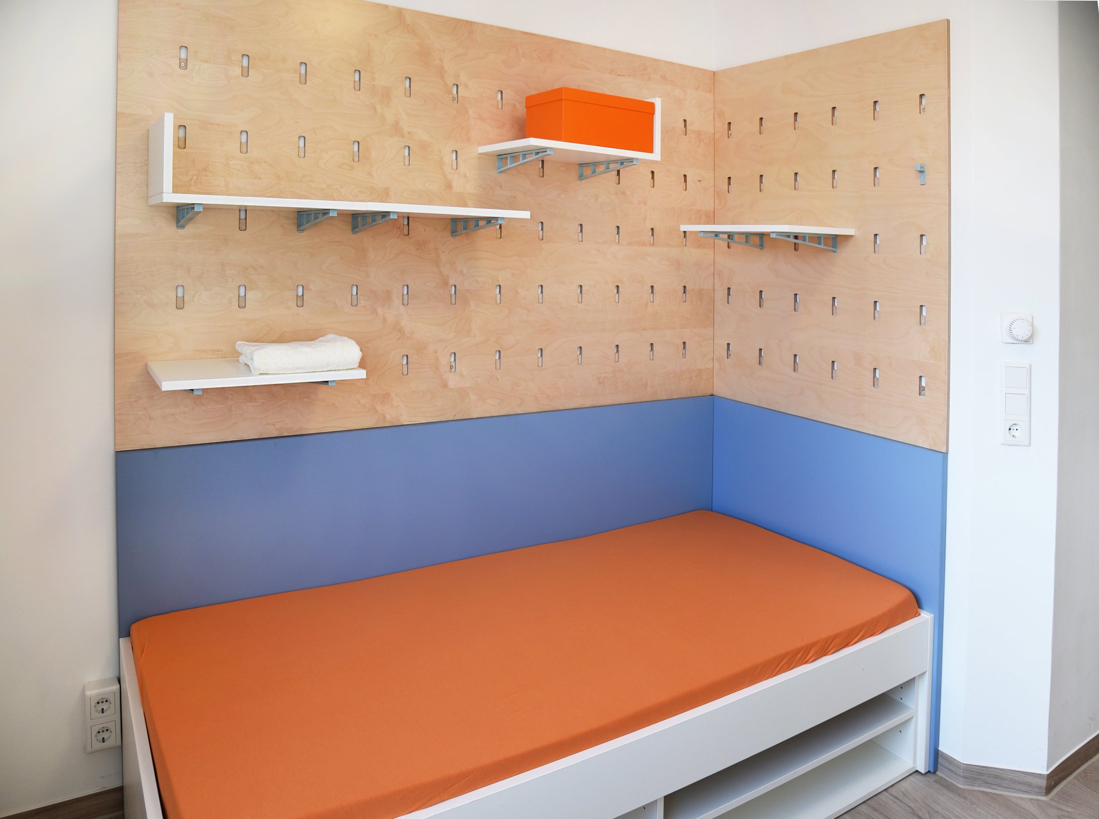 Modernes Zimmer eines Studierendenwohnheims in Paderborn-Peter_Hille_Weg_11 mit orangefarbenem Bett und Regalen.