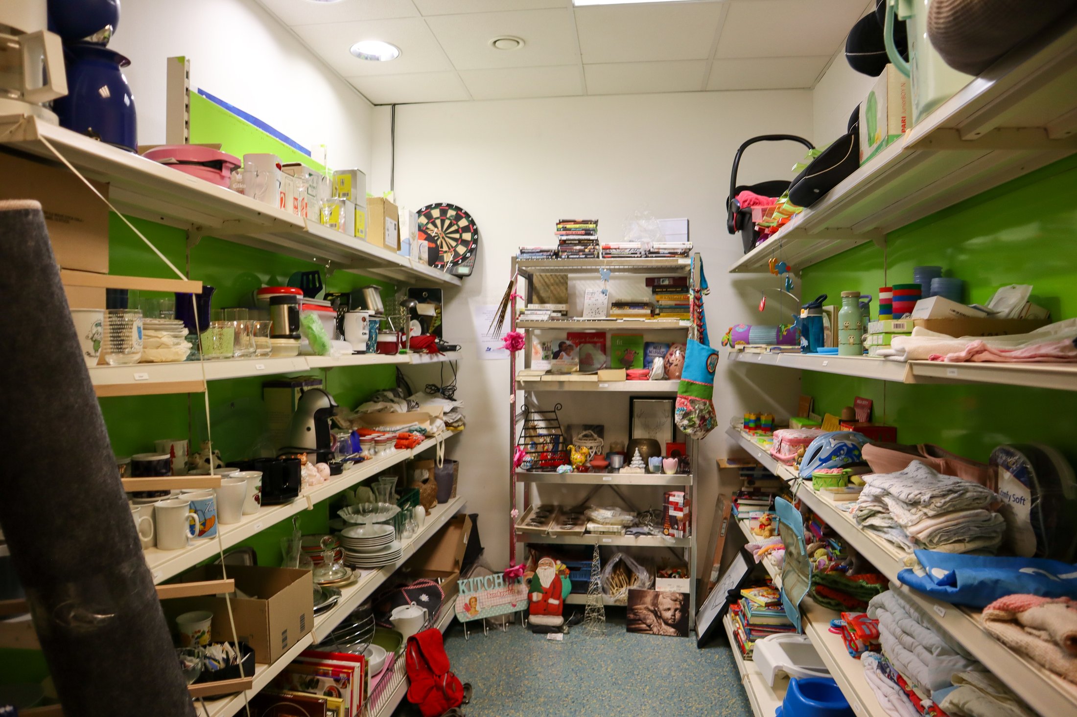 Regalreihen mit verschiedenen Haushaltswaren, Geschirr und Dekoration in einem hellen Raum mit grünen Wänden.