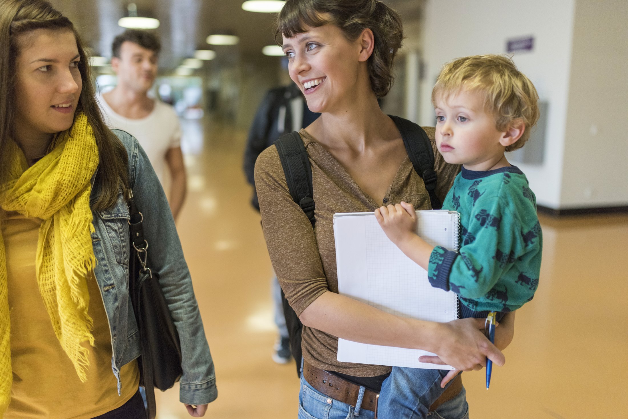 Zwei Frauen und ein Kind gehen in einem Flur. Eine Frau hält ein Notizbuch, während das Kind auf ihrem Arm sitzt.