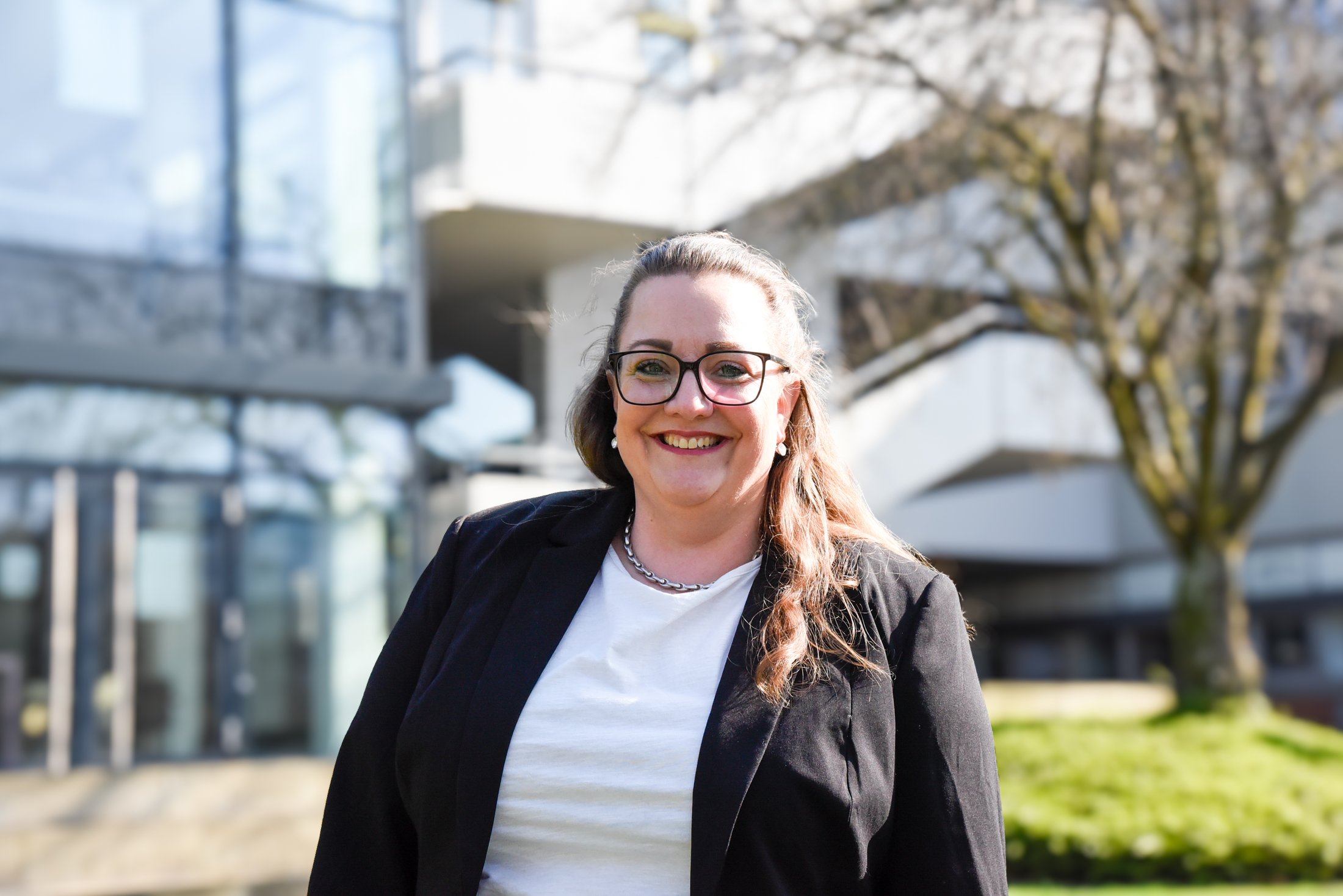 Eine Person steht im Freien vor einem modernen Gebäude mit großen Glasflächen und einem Baum im Hintergrund.