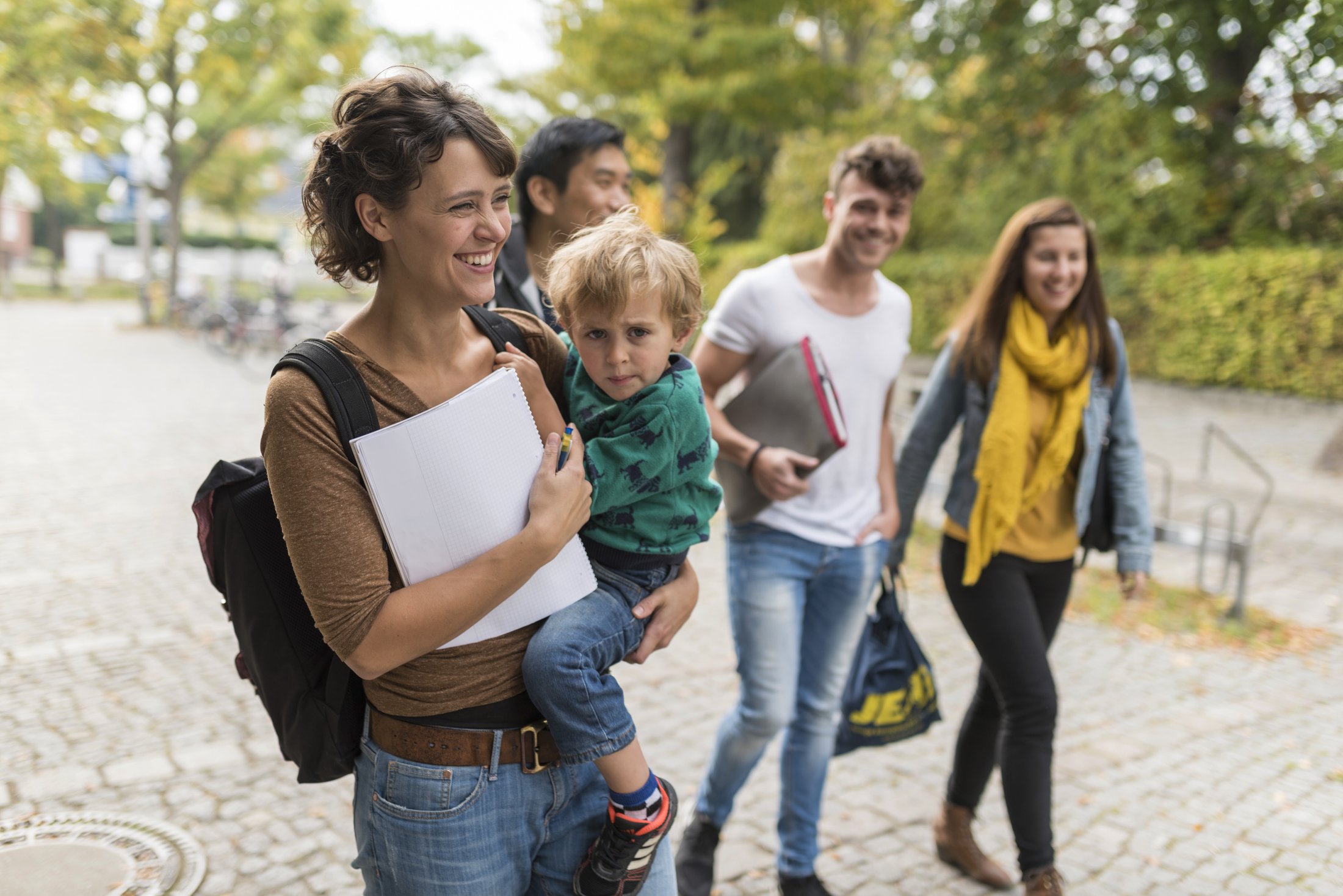 Gruppe von vier Personen auf einem gepflasterten Weg, eine Person trägt ein Kind, andere halten Unterlagen. Bäume im Hintergrund.