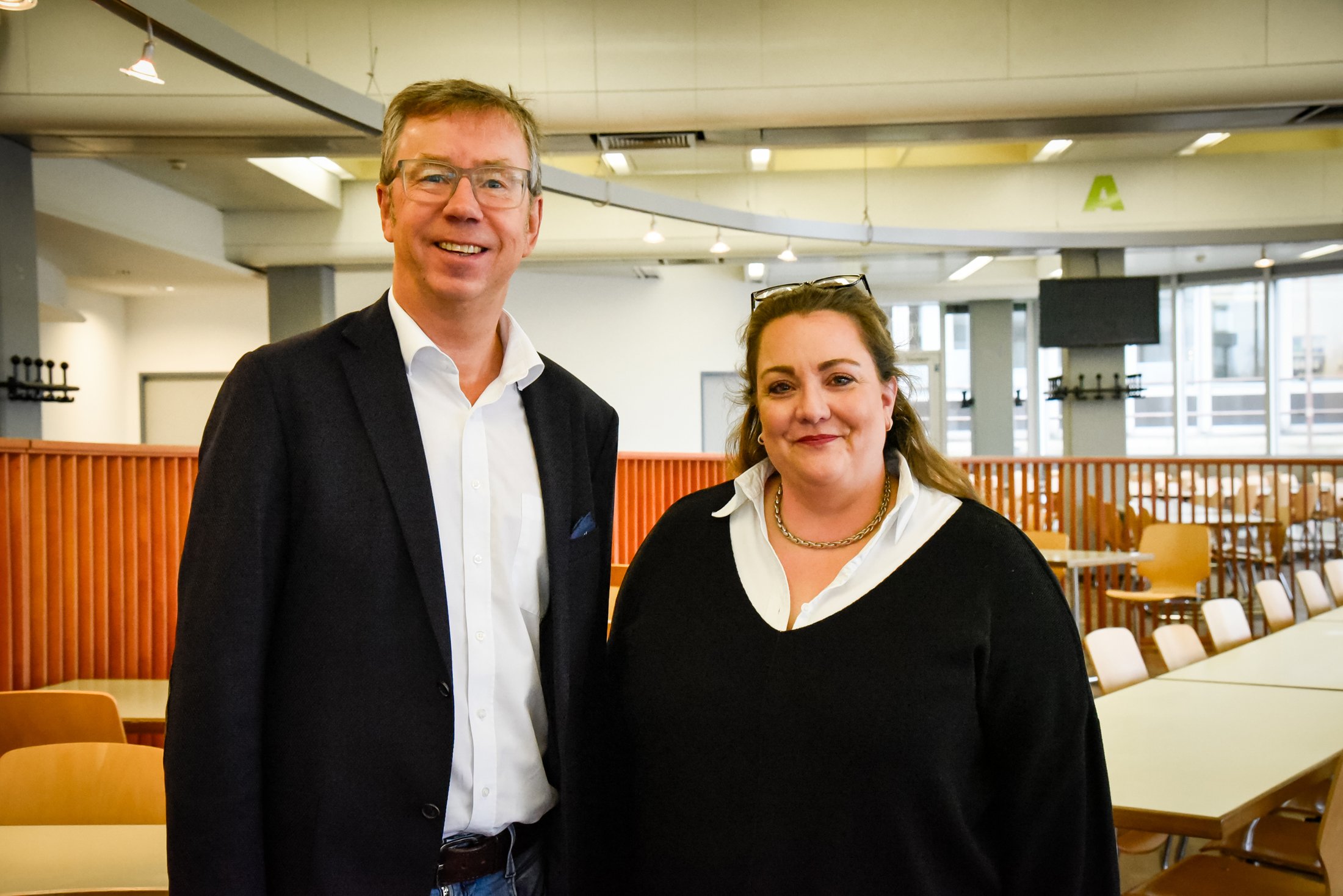 Zwei Personen lächeln in einem modernen Raum mit Tischen und Stühlen im Hintergrund. Helle, freundliche Atmosphäre.